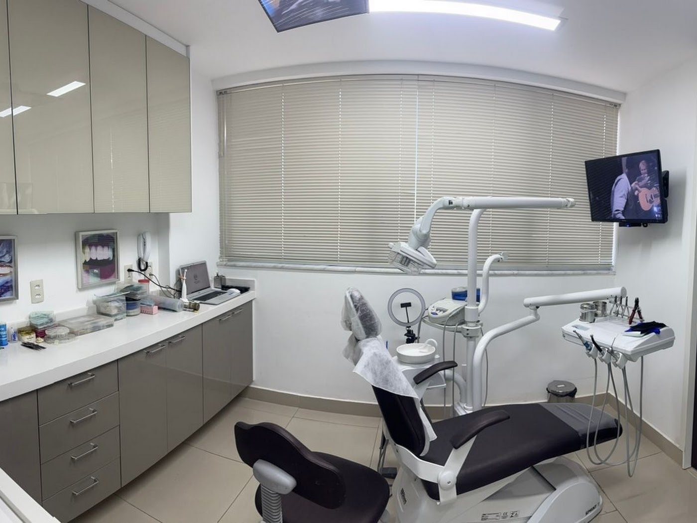 Sala de atendimento com a cadeira odontológica, armários claros e persiana na janela.